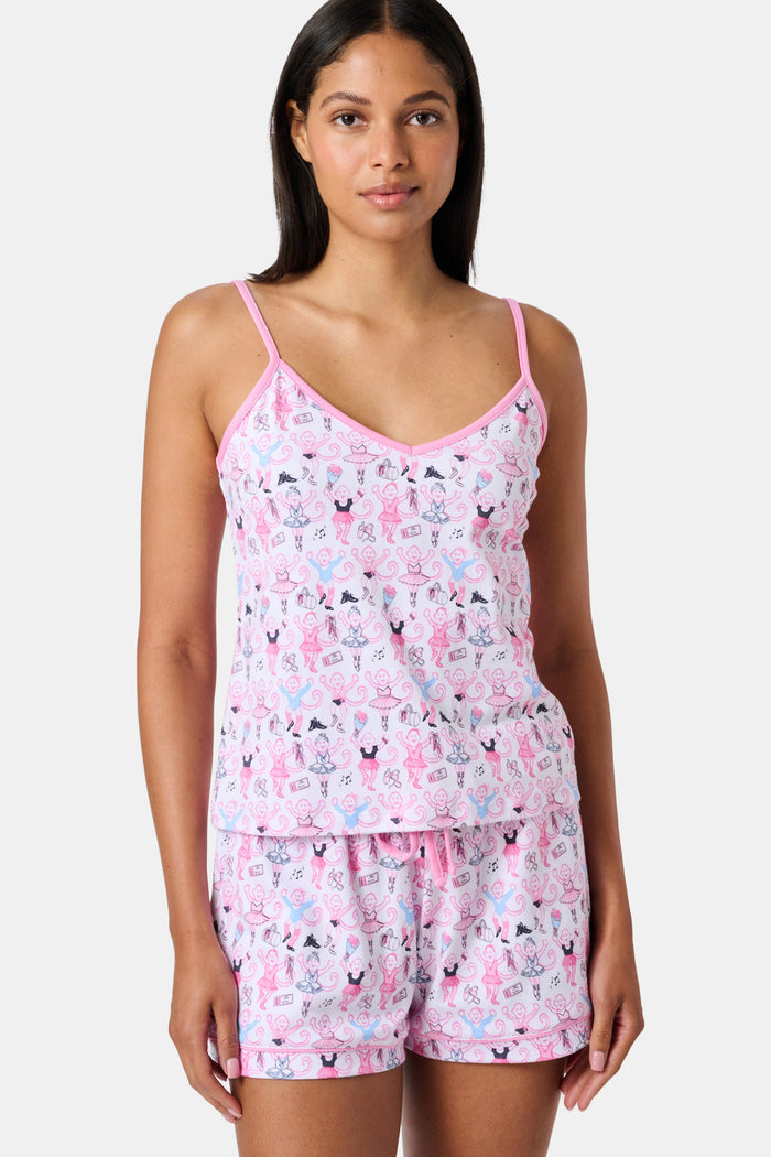 A woman wearing the Roller Rabbit Monkey Pirouette Mischa Set—pink and white cat-print Pima cotton loungewear—and holding a matching bag stands in front of white lockers, smiling slightly at the camera.