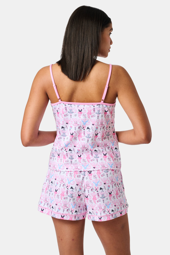 A woman wearing the Roller Rabbit Monkey Pirouette Mischa Set—pink and white cat-print Pima cotton loungewear—and holding a matching bag stands in front of white lockers, smiling slightly at the camera.