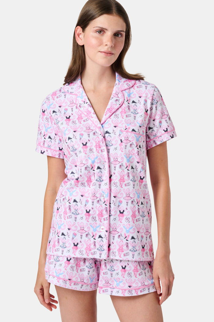 The Monkey Pirouette Polo Pajamas by Roller Rabbit feature a pink short-sleeve button-up top and matching shorts, adorned with a colorful and playful monkey, animal, and floral pattern on a white background.