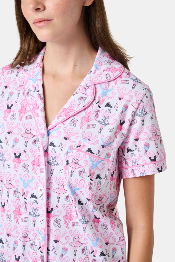 The Monkey Pirouette Polo Pajamas by Roller Rabbit feature a pink short-sleeve button-up top and matching shorts, adorned with a colorful and playful monkey, animal, and floral pattern on a white background.