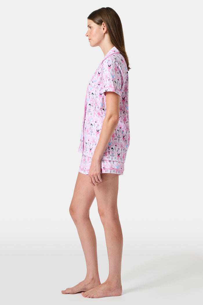 The Monkey Pirouette Polo Pajamas by Roller Rabbit feature a pink short-sleeve button-up top and matching shorts, adorned with a colorful and playful monkey, animal, and floral pattern on a white background.