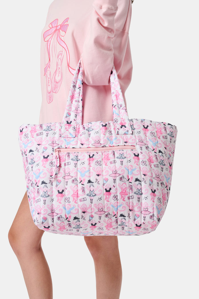Wearing matching pink and white floral shorts and top, a person stands by lockers holding the Roller Rabbit Monkey Pirouette Quilted Tote, paired with white socks and blue sneakers.