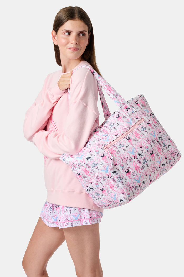 Wearing matching pink and white floral shorts and top, a person stands by lockers holding the Roller Rabbit Monkey Pirouette Quilted Tote, paired with white socks and blue sneakers.