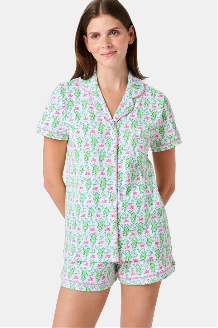 A woman wearing the Roller Rabbit Monkey Polo Pajamas, a short-sleeve button-up set with matching shorts featuring a green and pink floral pattern, stands against a plain white background.
