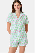 A woman wearing the Roller Rabbit Monkey Polo Pajamas, a short-sleeve button-up set with matching shorts featuring a green and pink floral pattern, stands against a plain white background.