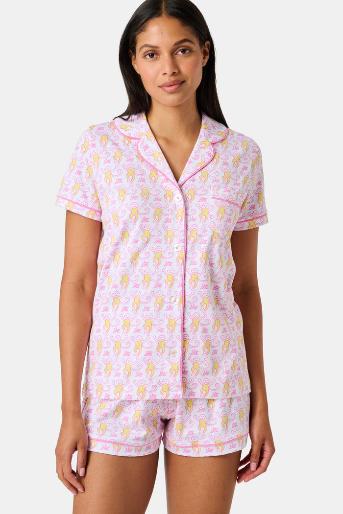 Introducing the Monkey Fiora Pajamas from Roller Rabbit—crafted from fabric adorned with a delightful pattern of pink and yellow monkeys with curled tails and smiling faces, interspersed with tiny leaves on a crisp white background. Perfect for adding playful charm to your sleepwear collection.