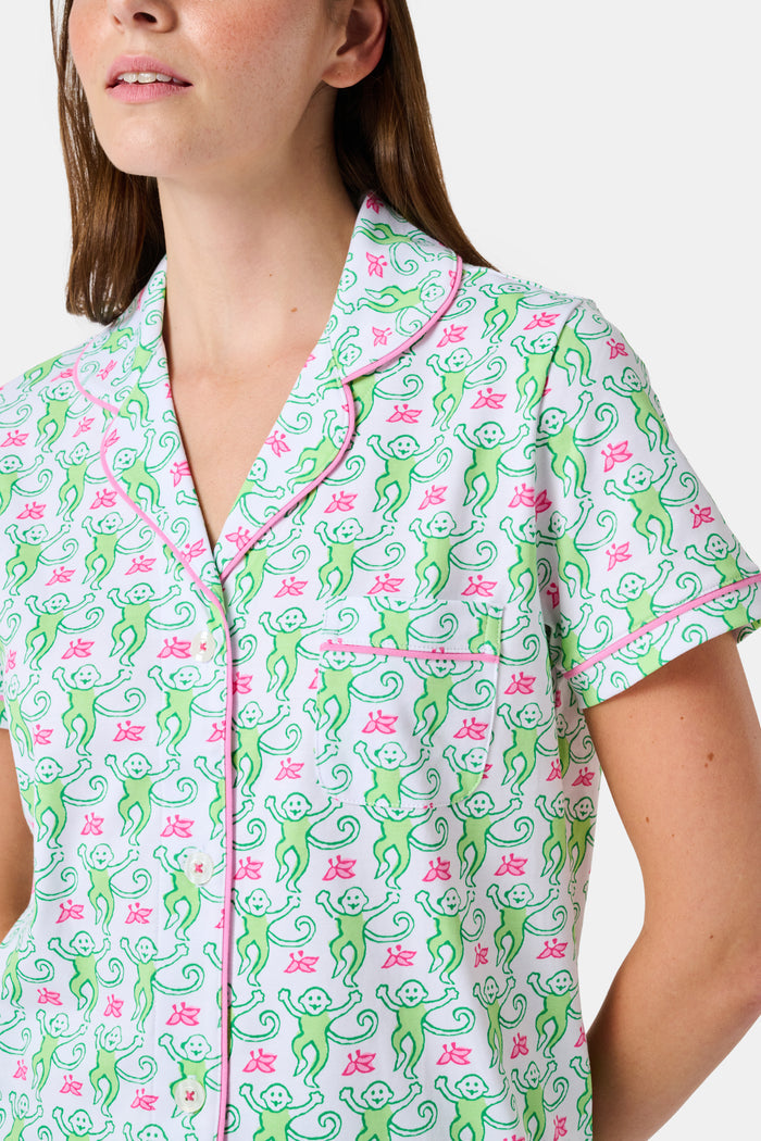 A woman wearing the Roller Rabbit Monkey Polo Pajamas, a short-sleeve button-up set with matching shorts featuring a green and pink floral pattern, stands against a plain white background.