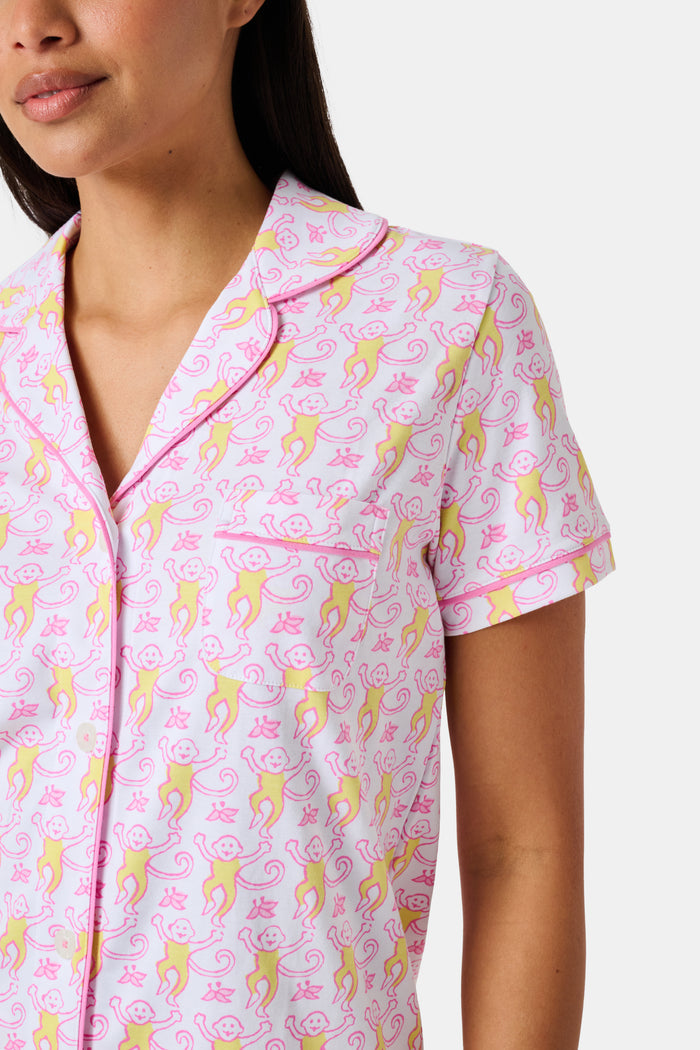 Introducing the Monkey Fiora Pajamas from Roller Rabbit—crafted from fabric adorned with a delightful pattern of pink and yellow monkeys with curled tails and smiling faces, interspersed with tiny leaves on a crisp white background. Perfect for adding playful charm to your sleepwear collection.