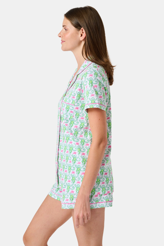 A woman wearing the Roller Rabbit Monkey Polo Pajamas, a short-sleeve button-up set with matching shorts featuring a green and pink floral pattern, stands against a plain white background.