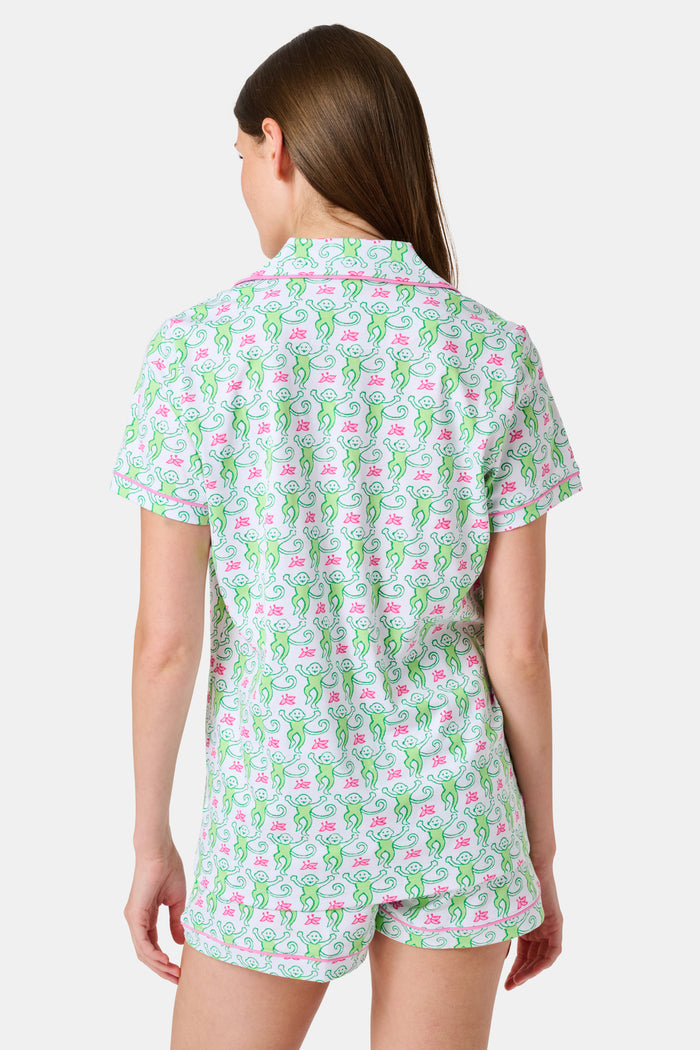 A woman wearing the Roller Rabbit Monkey Polo Pajamas, a short-sleeve button-up set with matching shorts featuring a green and pink floral pattern, stands against a plain white background.