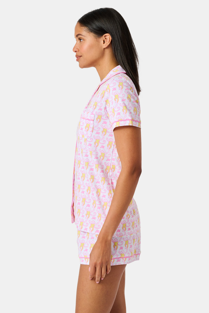 Introducing the Monkey Fiora Pajamas from Roller Rabbit—crafted from fabric adorned with a delightful pattern of pink and yellow monkeys with curled tails and smiling faces, interspersed with tiny leaves on a crisp white background. Perfect for adding playful charm to your sleepwear collection.