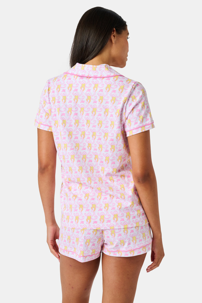 Introducing the Monkey Fiora Pajamas from Roller Rabbit—crafted from fabric adorned with a delightful pattern of pink and yellow monkeys with curled tails and smiling faces, interspersed with tiny leaves on a crisp white background. Perfect for adding playful charm to your sleepwear collection.