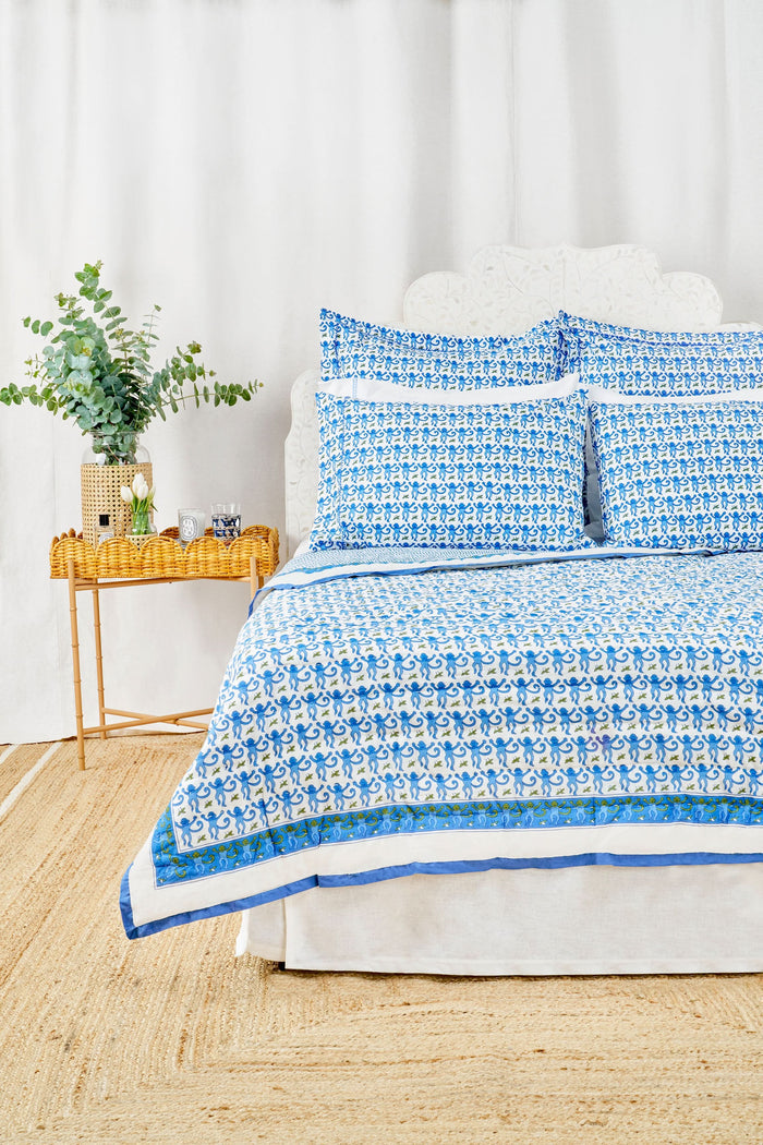 A neatly made bed features the Roller Rabbit Monkey Quilt with blue and white patterns and matching pillows, plus a lightweight throw. Next to it is a wicker nightstand with a vase of greenery, two glasses, and a woven rug covers the floor.