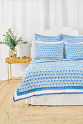 A neatly made bed features the Roller Rabbit Monkey Quilt with blue and white patterns and matching pillows, plus a lightweight throw. Next to it is a wicker nightstand with a vase of greenery, two glasses, and a woven rug covers the floor.
