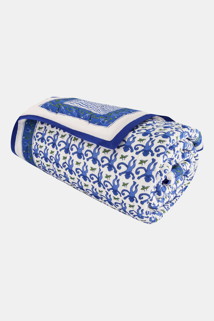 A neatly made bed features the Roller Rabbit Monkey Quilt with blue and white patterns and matching pillows, plus a lightweight throw. Next to it is a wicker nightstand with a vase of greenery, two glasses, and a woven rug covers the floor.