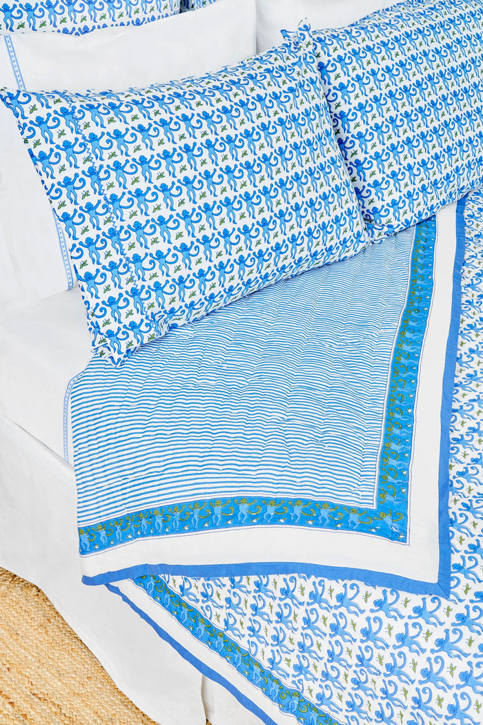 A neatly made bed features the Roller Rabbit Monkey Quilt with blue and white patterns and matching pillows, plus a lightweight throw. Next to it is a wicker nightstand with a vase of greenery, two glasses, and a woven rug covers the floor.