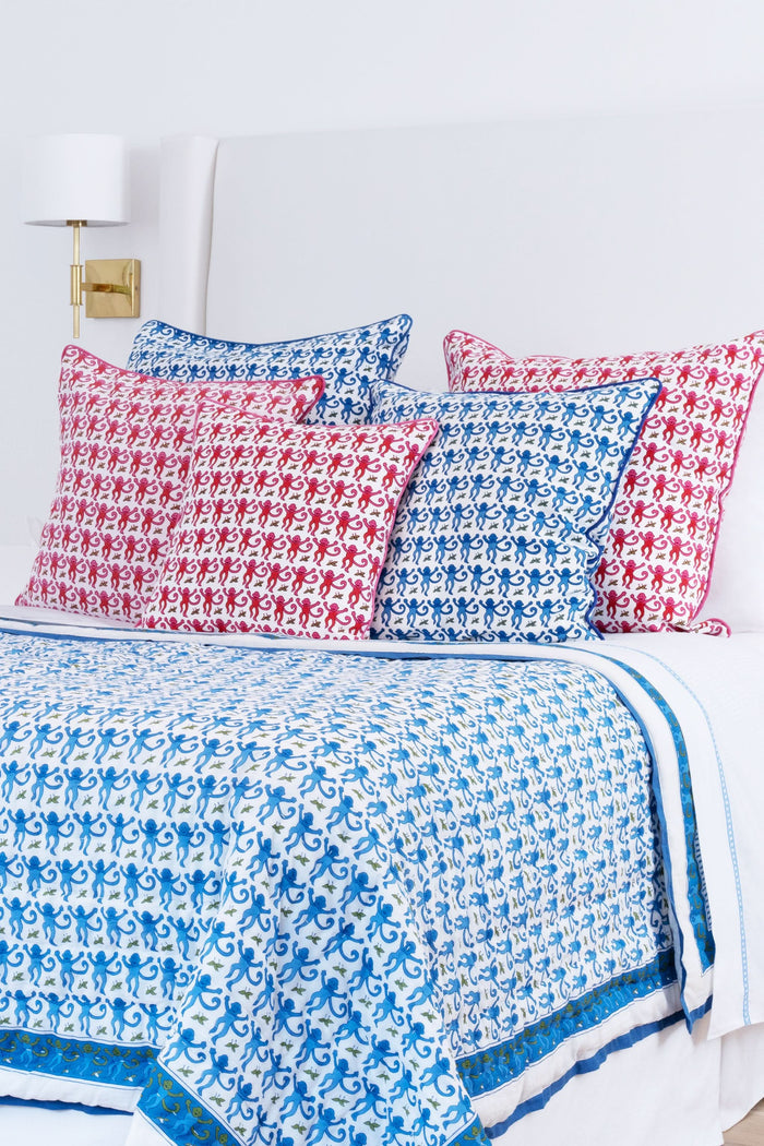 A neatly made bed features the Roller Rabbit Monkey Quilt with blue and white patterns and matching pillows, plus a lightweight throw. Next to it is a wicker nightstand with a vase of greenery, two glasses, and a woven rug covers the floor.