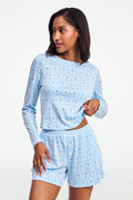 A person wearing the Roller Rabbit Monkey Rosebud Brooklyn Boxer pajama set, featuring a light blue menswear-inspired top and matching pima cotton shorts with a small floral print, poses against a plain white background.