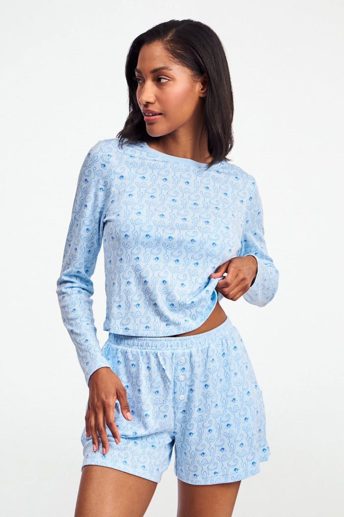 A person wearing the Roller Rabbit Monkey Rosebud Brooklyn Boxer pajama set, featuring a light blue menswear-inspired top and matching pima cotton shorts with a small floral print, poses against a plain white background.