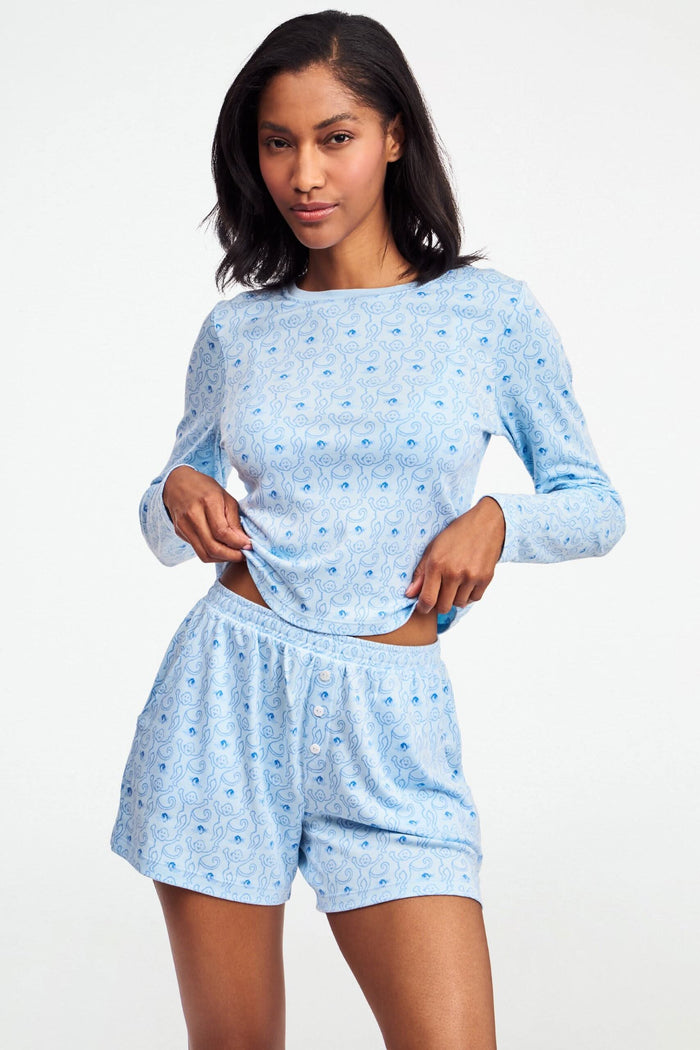 A person wearing the Roller Rabbit Monkey Rosebud Brooklyn Boxer pajama set, featuring a light blue menswear-inspired top and matching pima cotton shorts with a small floral print, poses against a plain white background.