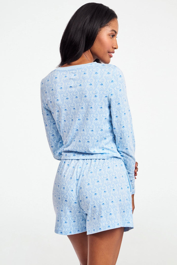A person wearing the Roller Rabbit Monkey Rosebud Brooklyn Boxer pajama set, featuring a light blue menswear-inspired top and matching pima cotton shorts with a small floral print, poses against a plain white background.