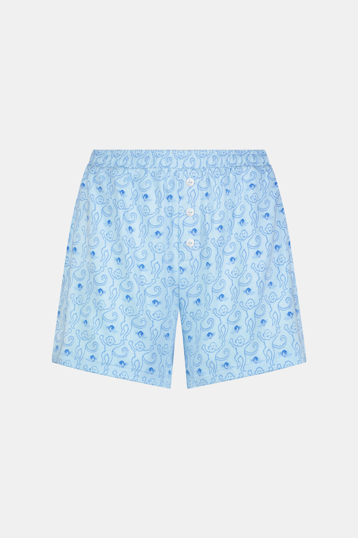 A person wearing the Roller Rabbit Monkey Rosebud Brooklyn Boxer pajama set, featuring a light blue menswear-inspired top and matching pima cotton shorts with a small floral print, poses against a plain white background.