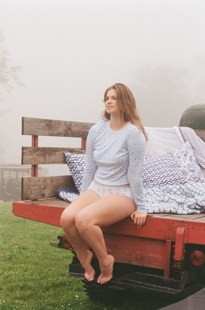 A woman with long brown hair wears the Roller Rabbit Monkey Rosebud Long Sleeve Baby Tee in light blue, featuring a subtle print, and matching shorts, while standing against a plain white background.