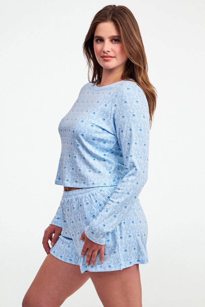 A woman with long brown hair wears the Roller Rabbit Monkey Rosebud Long Sleeve Baby Tee in light blue, featuring a subtle print, and matching shorts, while standing against a plain white background.