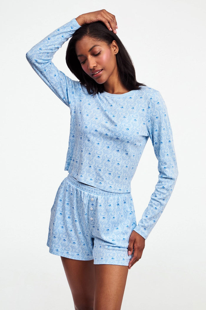 A woman with long brown hair wears the Roller Rabbit Monkey Rosebud Long Sleeve Baby Tee in light blue, featuring a subtle print, and matching shorts, while standing against a plain white background.