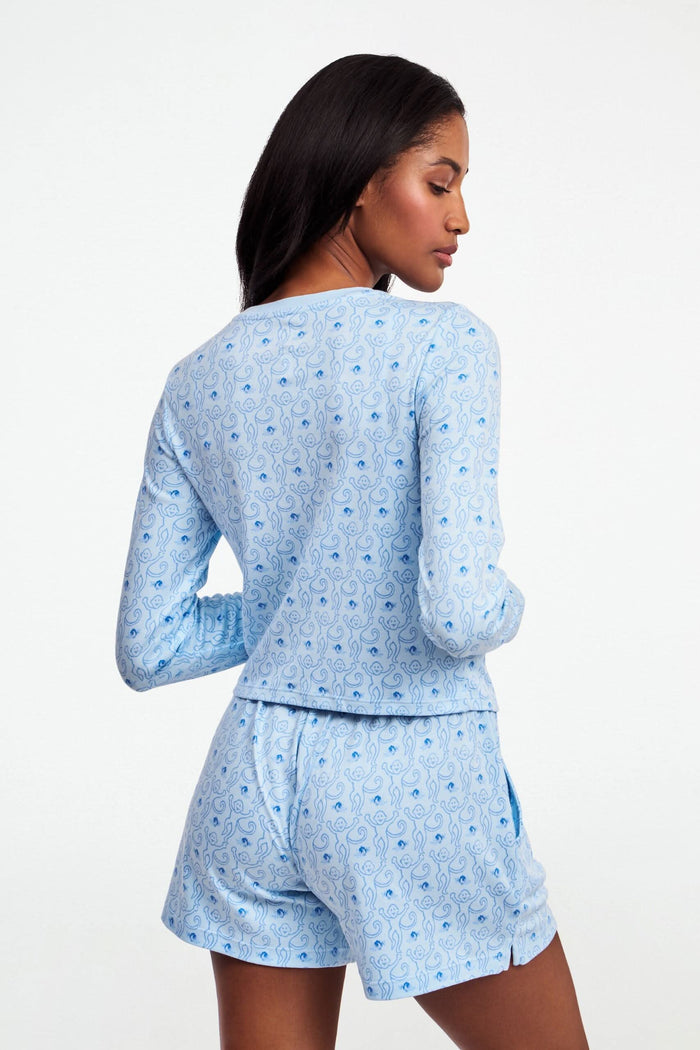 A woman with long brown hair wears the Roller Rabbit Monkey Rosebud Long Sleeve Baby Tee in light blue, featuring a subtle print, and matching shorts, while standing against a plain white background.