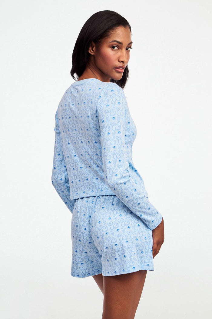 A woman with long brown hair wears the Roller Rabbit Monkey Rosebud Long Sleeve Baby Tee in light blue, featuring a subtle print, and matching shorts, while standing against a plain white background.