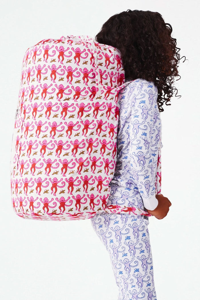 Wearing white and blue pajamas, a person carries the Roller Rabbit Monkey Sleepover Bag—an oversized bag featuring playful monkey prints—on their back. The bag is nearly as large as the person.