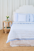 A bed features the Roller Rabbit Monkey/Hearts Reversible Quilt in light blue and white, with three matching pillows and a white headboard. Beside it, a round wicker side table holds flowers and books atop a woven rug.