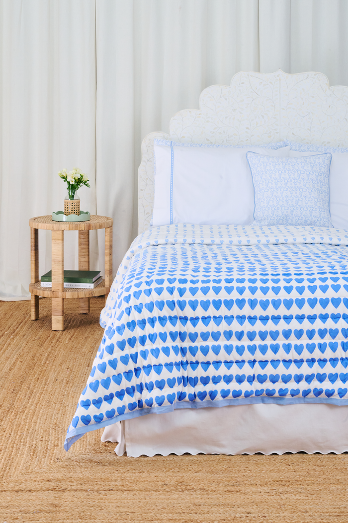 A bed features the Roller Rabbit Monkey/Hearts Reversible Quilt in light blue and white, with three matching pillows and a white headboard. Beside it, a round wicker side table holds flowers and books atop a woven rug.