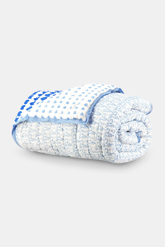 A bed features the Roller Rabbit Monkey/Hearts Reversible Quilt in light blue and white, with three matching pillows and a white headboard. Beside it, a round wicker side table holds flowers and books atop a woven rug.