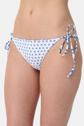 A person is shown from waist to mid-thigh wearing Roller Rabbit's Monkey/Hearts Serena Bikini Bottom, featuring blue heart motifs and blue-and-white side ties. The UPF 50+ swimwear combines stylish design with sun protection.