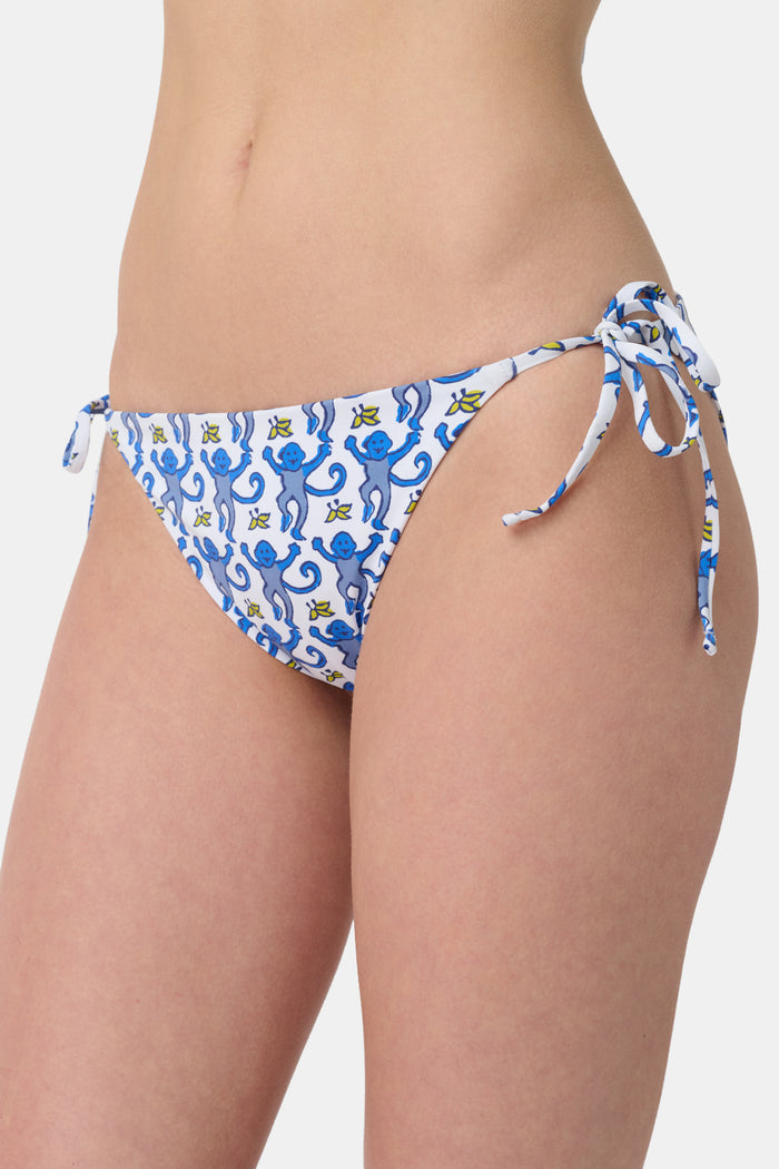 A person is shown from waist to mid-thigh wearing Roller Rabbit's Monkey/Hearts Serena Bikini Bottom, featuring blue heart motifs and blue-and-white side ties. The UPF 50+ swimwear combines stylish design with sun protection.