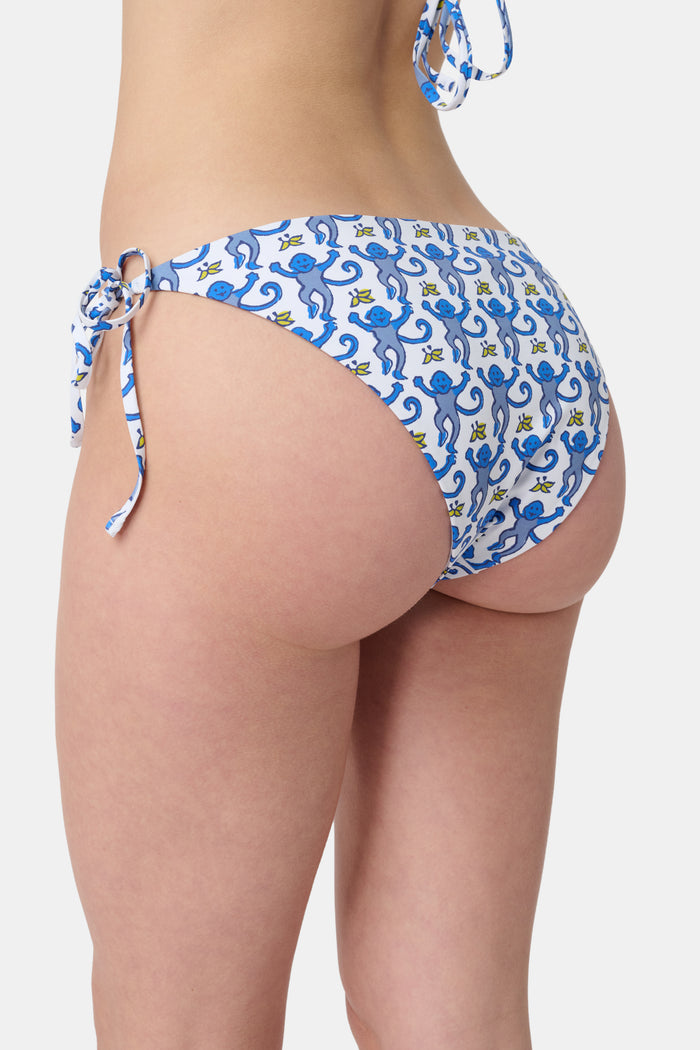 A person is shown from waist to mid-thigh wearing Roller Rabbit's Monkey/Hearts Serena Bikini Bottom, featuring blue heart motifs and blue-and-white side ties. The UPF 50+ swimwear combines stylish design with sun protection.
