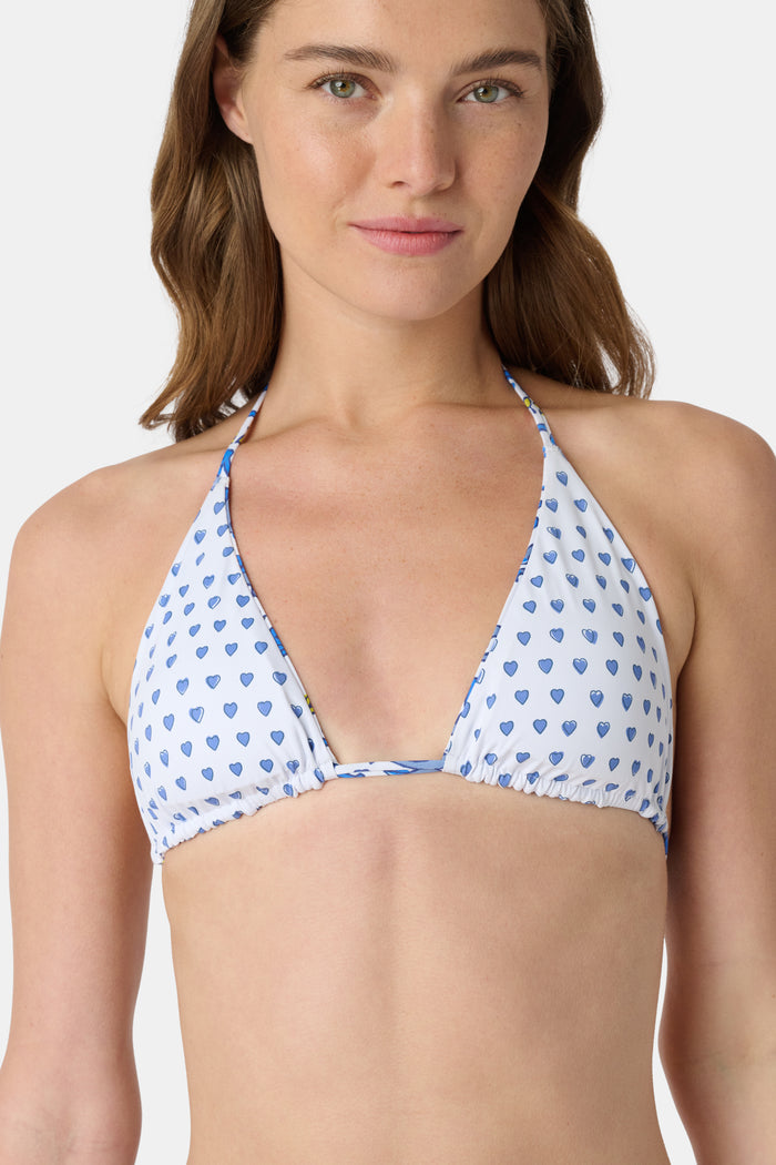 A woman wearing the Roller Rabbit Monkey/Hearts Serena Bikini Top, a white halter-style swim top with blue hearts, stands against a plain background and smiles softly.
