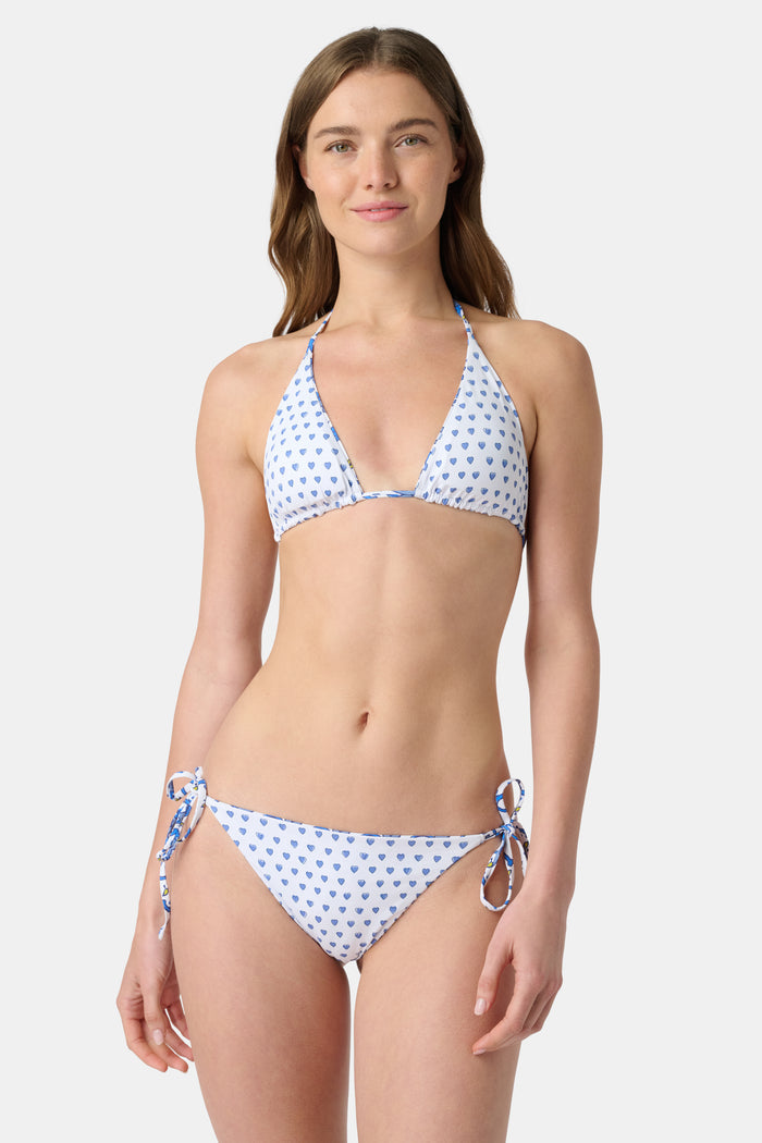 A woman wearing the Roller Rabbit Monkey/Hearts Serena Bikini Top, a white halter-style swim top with blue hearts, stands against a plain background and smiles softly.