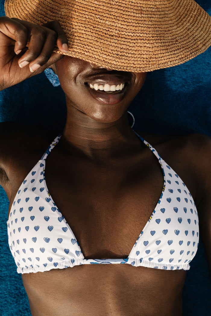 A woman wearing the Roller Rabbit Monkey/Hearts Serena Bikini Top, a white halter-style swim top with blue hearts, stands against a plain background and smiles softly.
