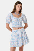 Two women in light blue and white skirts and blouses, one wearing the Roller Rabbit Naia Floral Stripe Fleet Skirt with a ruffle hemline, reach for books on a library shelf, standing side by side with their backs to the camera.