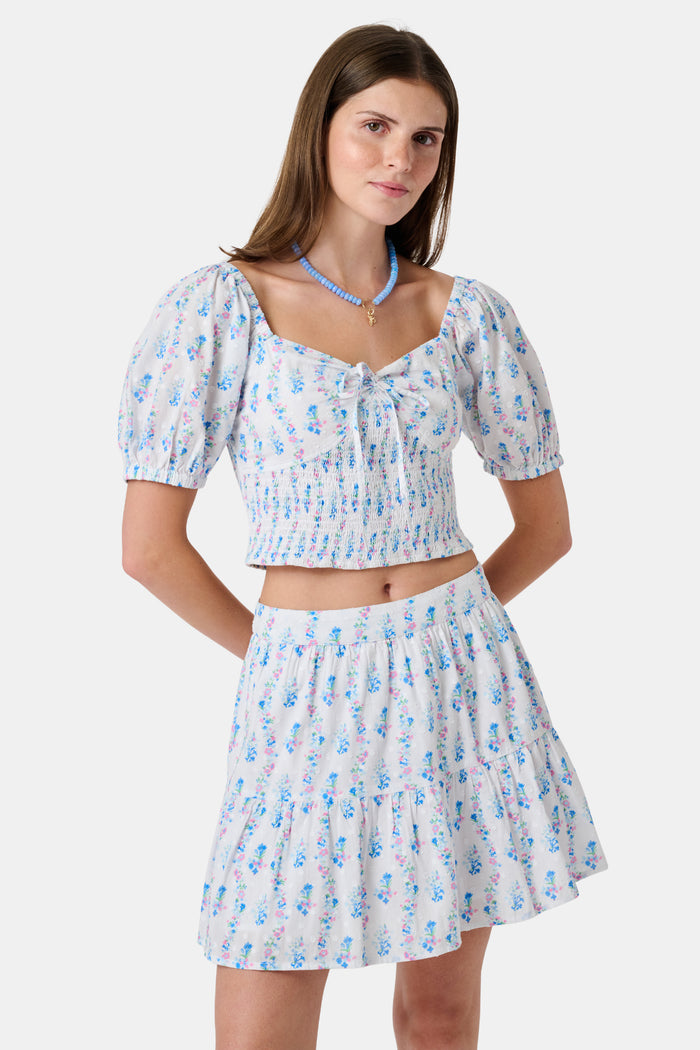 Two women in light blue and white skirts and blouses, one wearing the Roller Rabbit Naia Floral Stripe Fleet Skirt with a ruffle hemline, reach for books on a library shelf, standing side by side with their backs to the camera.