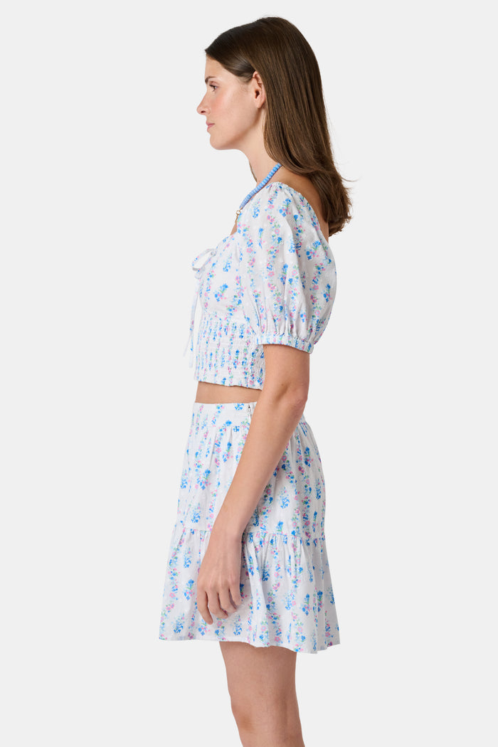 Two women in light blue and white skirts and blouses, one wearing the Roller Rabbit Naia Floral Stripe Fleet Skirt with a ruffle hemline, reach for books on a library shelf, standing side by side with their backs to the camera.