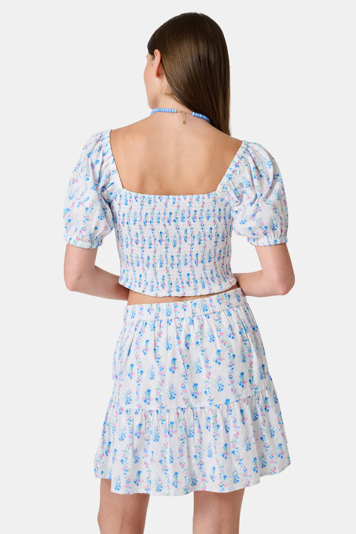 Two women in light blue and white skirts and blouses, one wearing the Roller Rabbit Naia Floral Stripe Fleet Skirt with a ruffle hemline, reach for books on a library shelf, standing side by side with their backs to the camera.