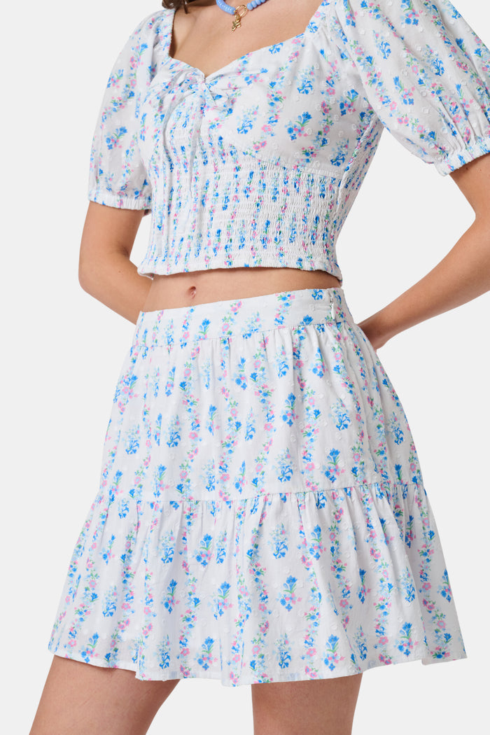 Four young girls stand and chat in front of white lockers, wearing pastel kids' dresses. One girl wears the Roller Rabbit Kids Naia Floral Stripe Alana Dress and holds a backpack and books, appearing to be in a school hallway. Another girl holds paper over her head.