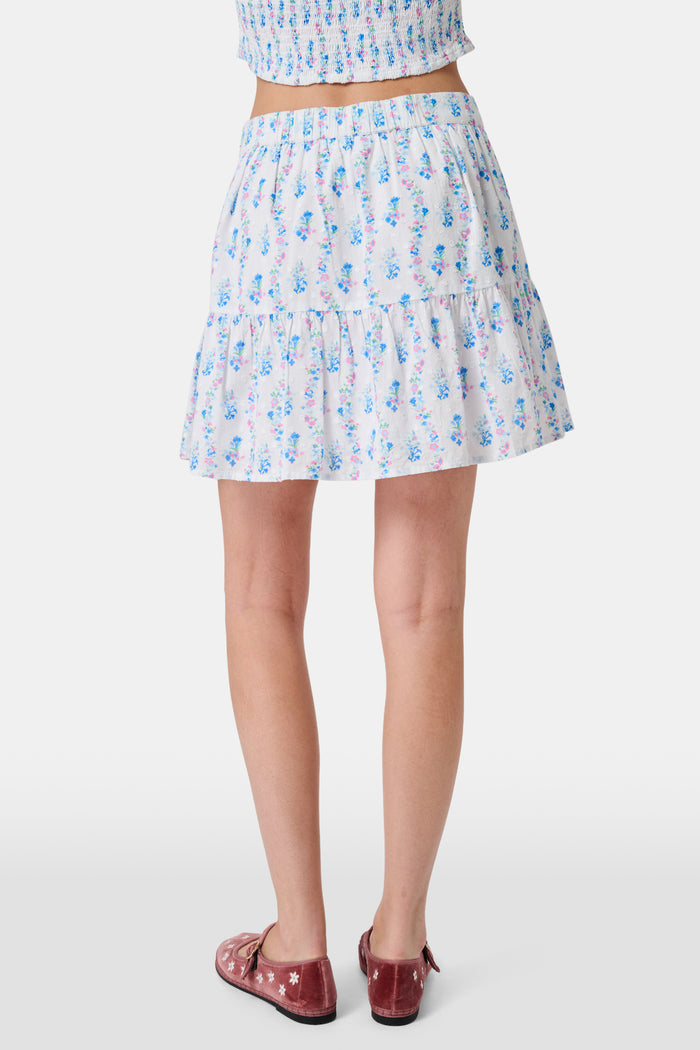 Four young girls stand and chat in front of white lockers, wearing pastel kids' dresses. One girl wears the Roller Rabbit Kids Naia Floral Stripe Alana Dress and holds a backpack and books, appearing to be in a school hallway. Another girl holds paper over her head.