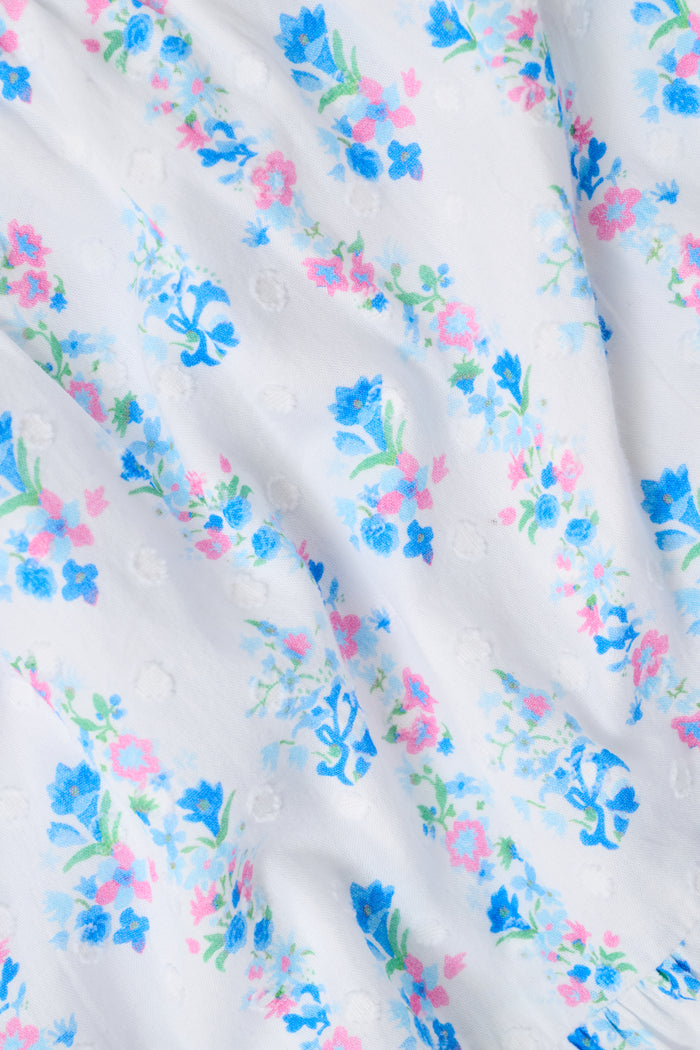 A woman with straight brown hair and a blue beaded necklace wears the Roller Rabbit Naia Floral Stripe Dovie Top—a light blue floral crop top with puff sleeves and a sweetheart neckline—paired with a matching skirt, standing against a plain white background.