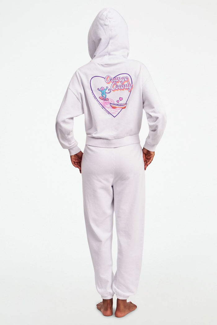 A person stands barefoot in Roller Rabbit O.C. Monkey Sweatpants and a matching hoodie, featuring colorful embroidered and printed graphics near the left pocket. The background is plain white.