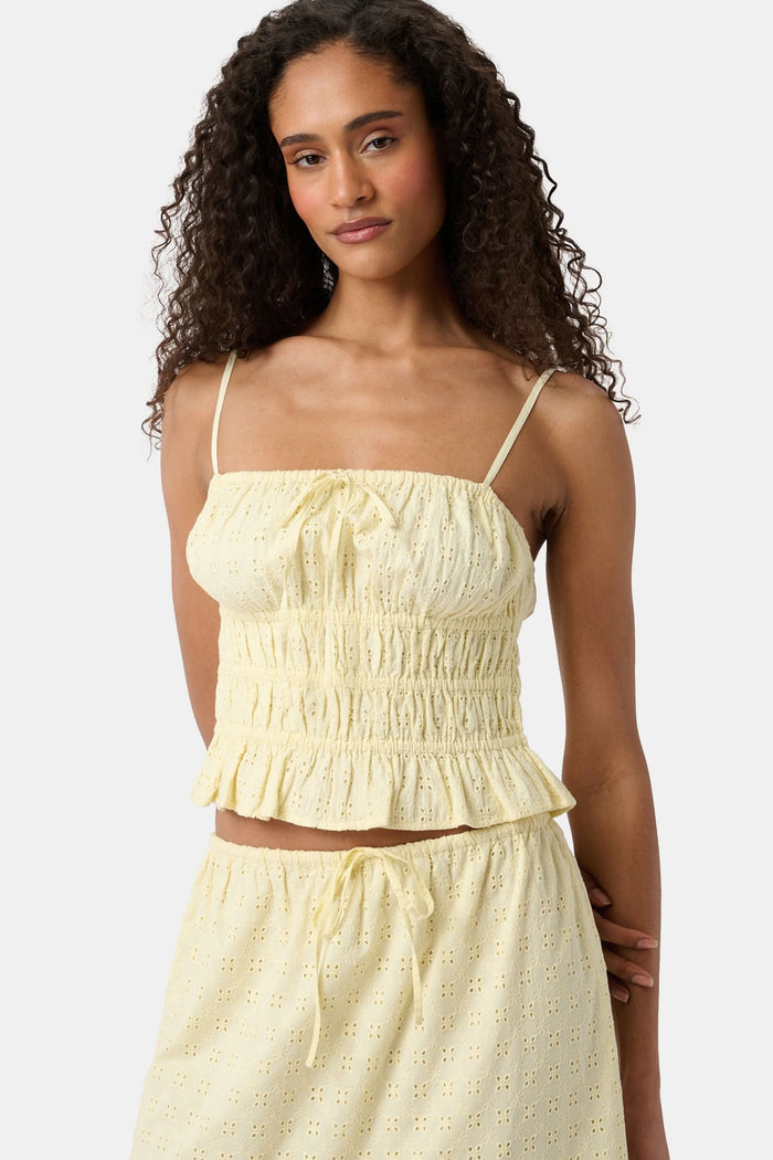 A woman with curly hair wears the Roller Rabbit Petal Eyelet Aurelia Top, a yellow cotton camisole with eyelet details and a ruffled hem, paired with a matching high-waisted skirt against a plain light background.